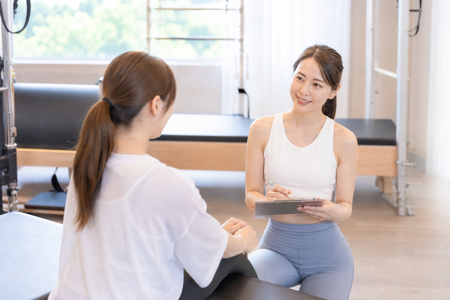 ピラティススタジオで女性がインストラクターと対面してカウンセリングを受けているシーン。初心者でも安心できるよう、インストラクターが親身に相談に乗っている様子。明るく開放的なスタジオ、信頼感のあるプロフ
