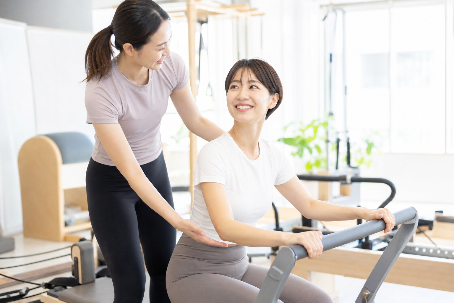 明るく親しみやすい雰囲気のピラティススタジオで、インストラクターが親切に女性の生徒を指導している様子。マンツーマンでマシンの使い方を教えており、生徒が笑顔で安心してトレーニングを楽しんでいる場面。清潔