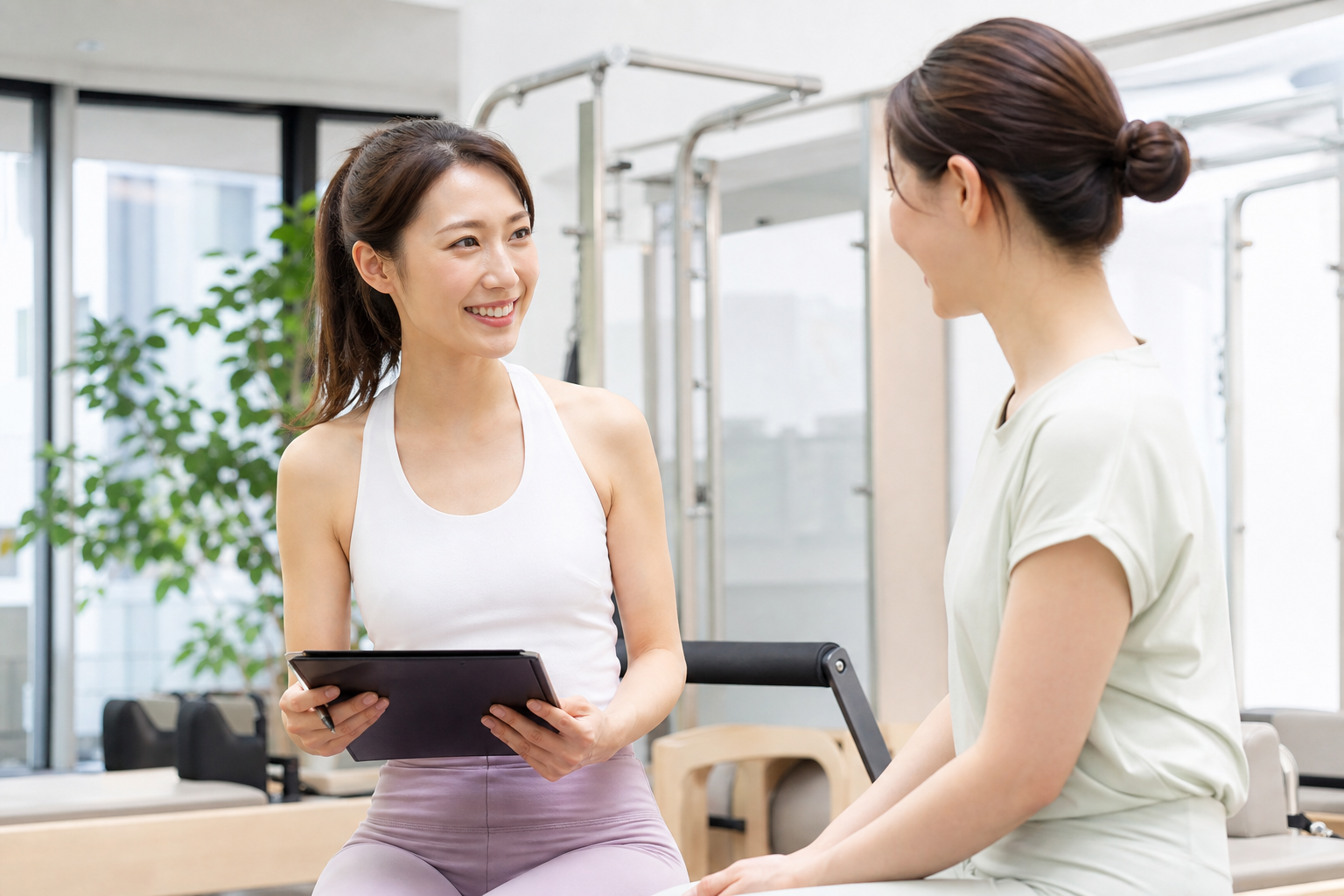 明るいスタジオでピラティスインストラクターが女性会員に笑顔でカウンセリングや指導を行っているシーン。実在の店舗ではない、親しみやすいインストラクターと初心者風の女性がコミュニケーションをとっているイメ