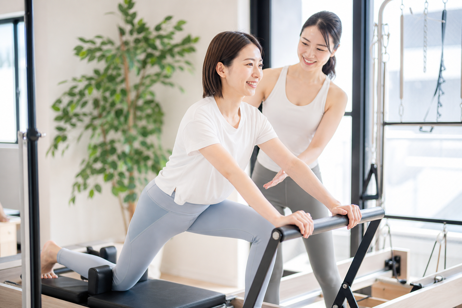 ピラティススタジオで女性がインストラクターの指導を受けながら、ピラティスマシン(リフォーマー)を使ってポーズをとっている様子。インストラクターは優しくサポートしており、明るくポジティブな雰囲気。初心者